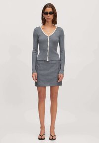 Grijze geribbelde cardigan met een V-hals en witte afwerking, gecombineerd met een aansluitende grijze minirok met zijzakken. Model draagt zwarte zonnebril.