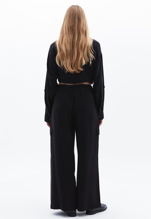 Femme aux longs cheveux blonds portant un haut noir à manches longues et un pantalon large, debout de dos sur fond blanc.