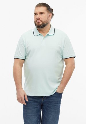 Mężczyzna z brodą i związanymi włosami nosi jasnoniebieską koszulkę polo z ciemnym obszyciem i niebieskie dżinsy, stoi z jedną ręką w kieszeni, patrząc na bok.