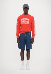 Czerwona bluza z nadrukiem "Aries", dżinsowe szorty, białe skarpetki i białe sneakersy. Model nosi szary czapkę. Prosty, casualowy strój.