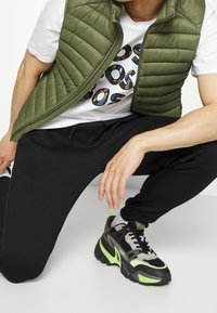 Chaleco acolchado verde sobre una camiseta blanca con un logo multicolor, combinado con pantalones jogger negros y zapatillas deportivas negras con detalles verdes.