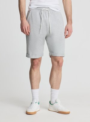 Mężczyzna w jasnoszortach z sznurkiem, białych skarpetkach za kostkę i białych trampkach z zielonymi akcentami, stojący na jednolitym tle.