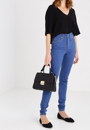 Woman wearing a black V-neck top, blue high-waist skinny jeans, black flats, holding a black handbag with gold clasp.