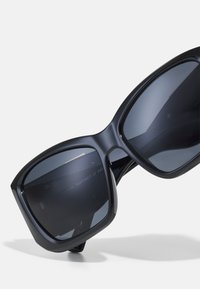 Occhiali da sole neri con forma a occhio di gatto oversize. Presentano lenti scure e riflettenti e montature in plastica liscia. Hardware minimale visibile.
