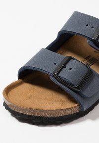 Blå syntetisk sandal med en texturerad yta, två justerbara remmar med svarta spännen samt en brun korksula och svart gummisål.
