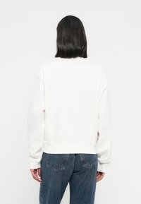 Άσπρο βαμβακερό φούτερ με άνετη εφαρμογή, λα ιμόκοψη τύπου crew και χαμηλωμένους ώμους. Συνδυάζεται με σκούρο μπλε τζιν παντελόνι. Χωρίς ορατά λογότυπα ή σχέδια.