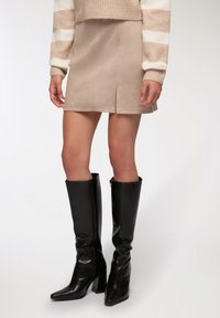 Gonna mini in suede beige con spacco frontale, abbinata a stivali alti in pelle nera con tacco, dalla punta affusolata e parte superiore arrotondata.