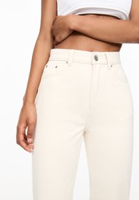 Jean taille haute beige clair en denim, avec une fermeture à boutons, cinq poches et des détails matériels subtils.