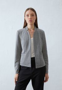 Giacca grigia a spina di pesce con colletto rialzato, slim fit e tasche laterali. Indossata su una maglietta bianca, abbinata a pantaloni neri. Texture liscia.