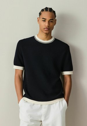 Junger Mann mit geflochtenem Haar, der ein schwarzes kurzärmeliges Strickshirt mit weißem Besatz und weiße Hose trägt, steht mit den Händen in den Taschen.