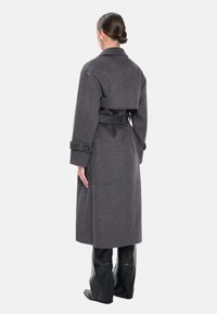 Grå uldfrakke med bælte i taljen, bred krave og knaplukning ved ærmekanterne, båret over sorte, blanke bukser. Glat tekstur og lige silhuet.