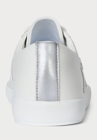Sneaker blanche avec une tige en cuir lisse, accent métallique argenté au niveau du talon, semelle plate et détails de couture contrastés.