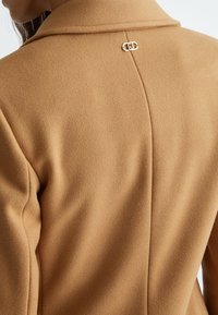 Cappotto in misto lana marrone con una finitura tessurizzata, dotato di colletto a punta e dettaglio con chiusura in tonalità oro sul retro. Vestibilità sartoriale con linee di cucitura.