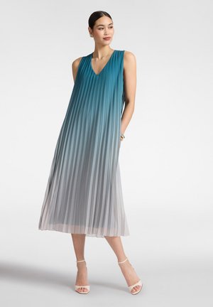 Frau trägt ärmelloses, plissiertes Kleid mit V-Ausschnitt und Ombre-Färbung von Blau bis Hellgrau, posiert mit einer Hand in der Tasche und beigen Knöchelriemen-Absatzschuhen.