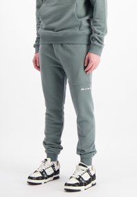 Pantalons de survêtement vert clair avec des poignets élastiques et des poches avant, arborant un logo blanc "BLCK" au bas de la jambe. Portés avec des baskets à motifs.