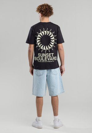 Camiseta negra con un gráfico del sol y el texto "SUNSET BOULEVARD" en crema. Combinada con pantalones cortos de mezclilla claros y zapatillas blancas.