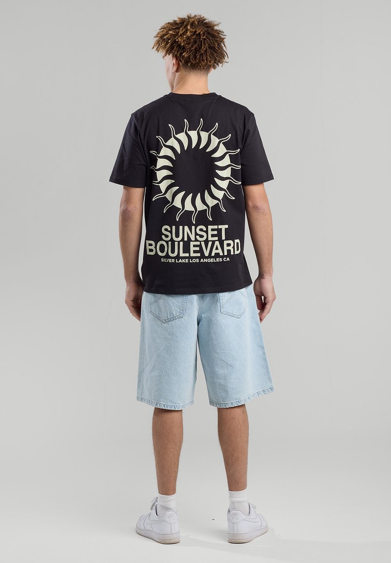 Black t-shirt with a sun graphic and "SUNSET BOULEVARD" text in cream. Paired with light denim shorts and white sneakers.