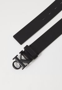 Cintura in pelle nera con design a nastro piatto e fibbia nera opaca a forma di "CK". Presenta più fori di regolazione lungo il nastro.