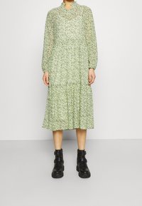 Vihreä kukkakuosis midi-mekko pitkillä hihoilla, korkealla pääntiellä, kerroksellisella helman ja puolipaineellisella rakenteella, yhdistettyinä mustiin nilkkabootseihin.