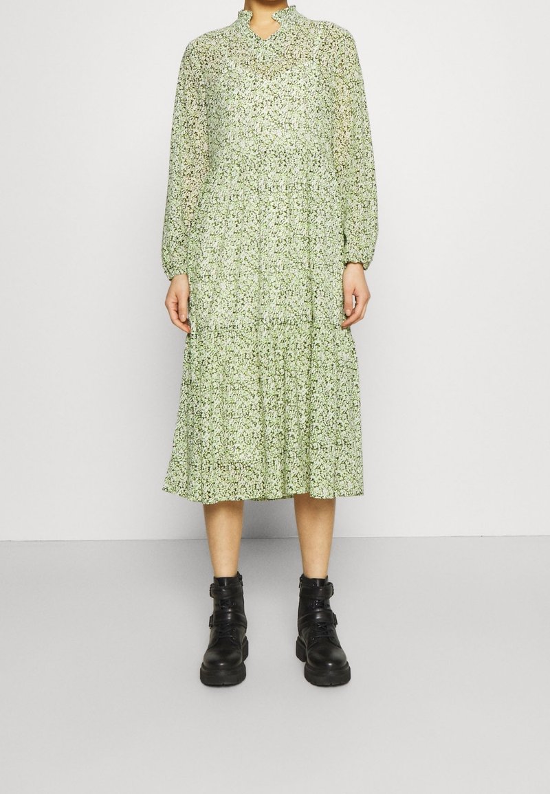 Zelené květinové midi šaty s dlouhými rukávy, vysokým výstřihem, vrstvenou sukní a poloprůsvitnou texturou, doplněné černými kotníkovými botami.