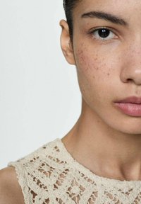 Beige lace top featuring a textured crochet pattern, worn by a person with a smooth complexion and neutral makeup, focusing on facial features.