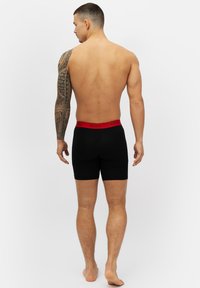 Svarta boxerkalsonger med en röd elastisk midjeband; slät textur och åtsittande design. Visar en manlig modell med en synlig tatuering på armen.