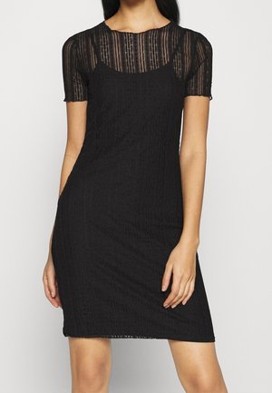 Kvinde iført en sort kortærmet kjole med gennemsigtig blonder øverste del og ærmer, stående mod en ensartet lys baggrund.