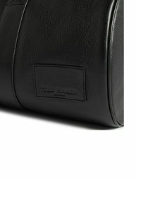 Sac en cuir noir à texture lisse, forme courbée, avec un patch rectangulaire marqué en relief du nom "TED BAKER". Détails de coutures apparents.