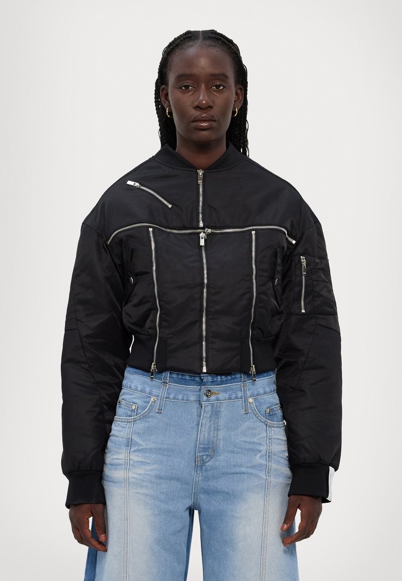 Zwarte cropped bomberjack met zilveren ritsdetails, voorzien van lange mouwen en een staande kraag, gecombineerd met lichtblauwe jeans.