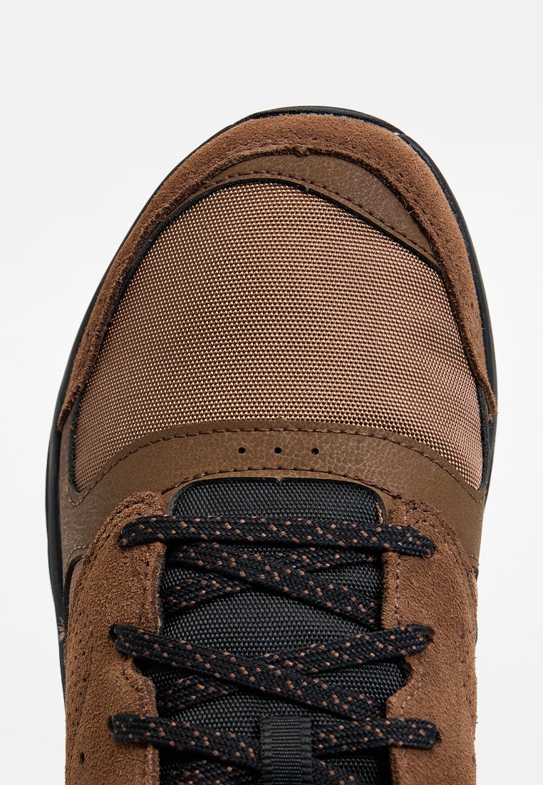 Zapatilla marrón con una parte superior de cuero texturizado y malla, que presenta cordones oscuros y una suela de goma negra. Los detalles de costura acentúan el diseño.