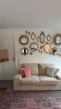 Beige stoffen bank met twee kussens, één gestreept, één met textuur. De muur toont verschillende sierlijke spiegels in gouden en zilveren kaders. Kleurige vloerkleed eronder.