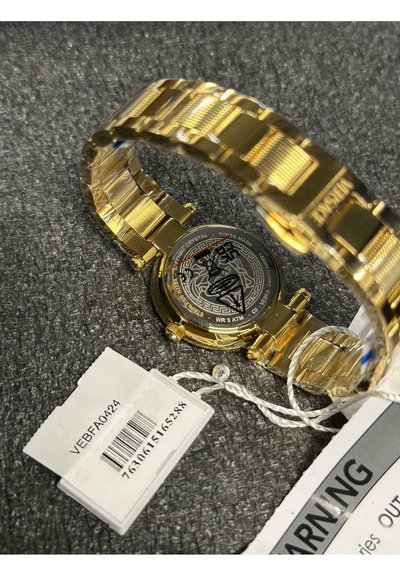 Guldfarvet metal wristwatch med en mønstret sort urskive, udstillingsetui og et poleret ledarmbånd. Inkluderer et mærke med en stregkode.