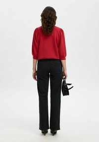 Donna con i capelli ricci, indossa una blusa rossa, jeans neri, tacchi neri e tiene una borsa nera, in piedi contro uno sfondo semplice.