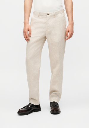 Man die lichtbeige op maat gemaakte broek en donkerbruine leren loafers draagt, staand tegen een effen witte achtergrond.