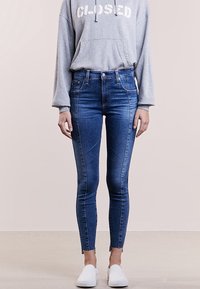 Úzké džíny v tmavě modrém denimu s vysokým pasem, s jemným vyblednutím, pěti kapsami a surovým lemem na kotníku, nošené s šedou mikinou.