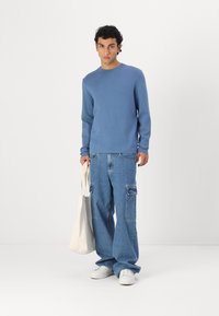 Zils garās piedurknes džemperis, brīvi denim cargo bikses ar sānu kabatām un baltas kedas. Modelis tur kanvas somu.