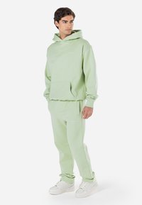 Lys grønn hettegenser og matchende joggebukser med lommer foran og logo. Laget av mykt stoff, har elastiske mansjetter og midjebånd.