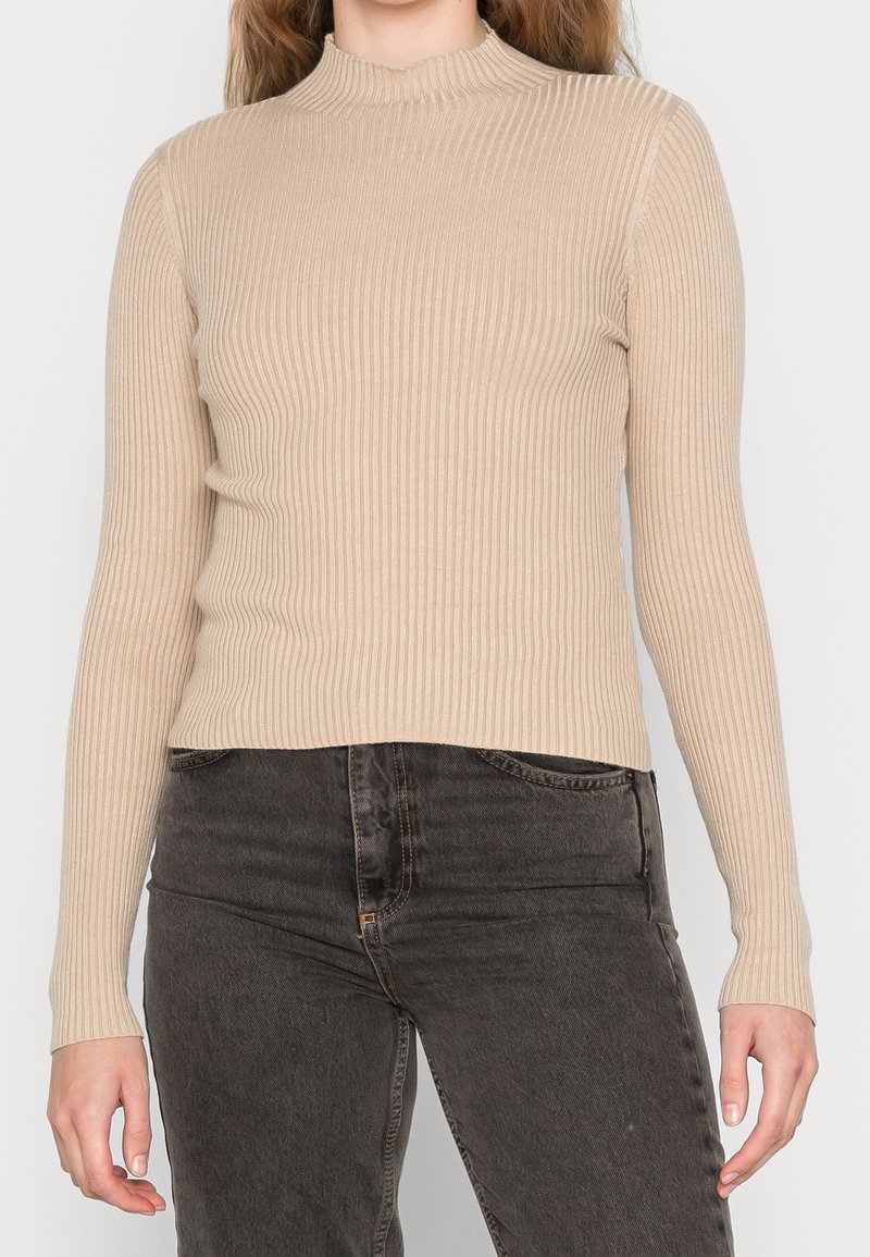 Femme portant un haut beige à manches longues côtelé et un jean gris foncé taille haute, debout avec les bras détendus le long du corps.