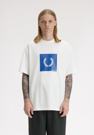 Jeune personne aux cheveux mi-longs portant un t-shirt blanc oversize avec un logo de couronne de laurier bleu et un pantalon noir, debout devant un fond blanc.