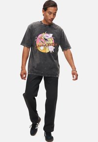 Svart bleknad bomullströja med en färgglad cirkulär grafik som har figurer och texten "Ko och Kyckling." Kombinerad med svarta byxor och sneakers.