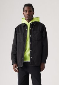 Zwart denimjack met twee voorzakken en zilveren drukknoopsluitingen, gelaagd over een felgroene hoodie.