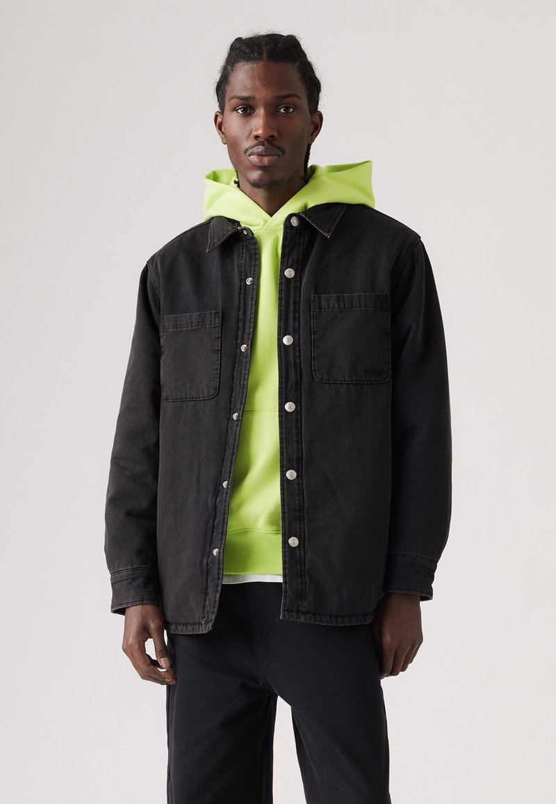 Zwart denimjack met twee voorzakken en zilveren drukknoopsluitingen, gelaagd over een felgroene hoodie.