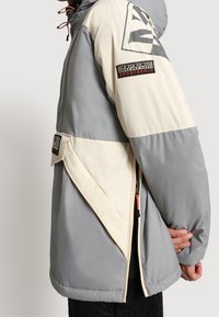 Grå och gräddfärgad färgblockerad jacka med huva, som har en triangulär logotyp, fickor med dragkedja och kontrasterande orangea detaljer.