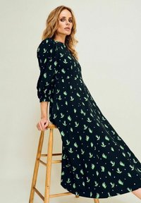 Musta mekko, jossa on vihreä lehtikuvio, puffihihat ja keskipitkä helma. Sileä tekstuuri, löysä istuvuus ja hienovaraiset valkoiset yksityiskohdat kauttaaltaan.