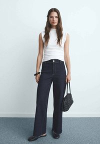 Witte mouwloze top, high-waisted donkere denim wijde broek met afgesneden zoom, zwarte gevlochten handtas, zwarte instapschoenen, neutrale achtergrond.