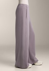 Světlé fialové široké kalhoty s texturovaným pasem a bočními kapsami, vyrobené z měkké látky. V kombinaci s bílými sportovními botami.