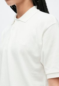 Vit pikétröja i texturerat tyg med klassisk krage och en placket med tre knappar. Diskret broderad logotyp nära bröstet.