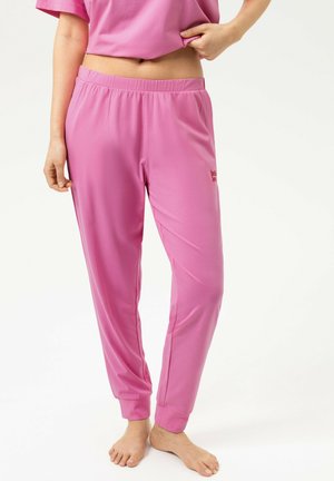 Persona che indossa pantaloni jogger rosa con vita elastica e top corto abbinato, in piedi a piedi nudi su sfondo bianco.