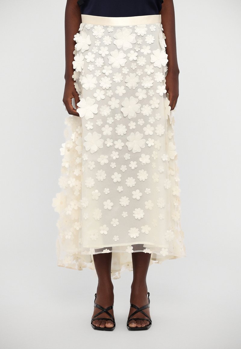 Jupe midi blanche translucide avec applications florales 3D superposées, portée avec des sandales à talons noires à brides.