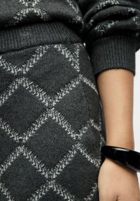 Pull en tricot gris avec un motif en losanges en gris clair. Doté de poignets côtelés et d'une coupe slim, associé à un bracelet noir au poignet.